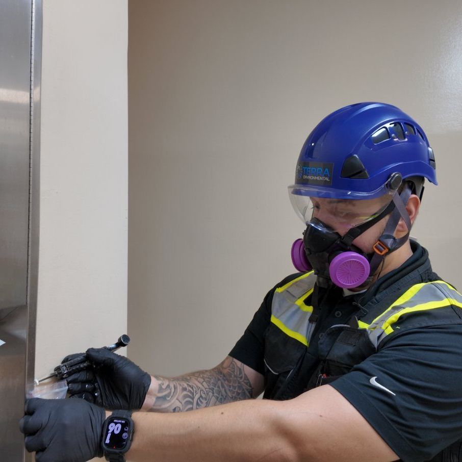 TERRA technician in respirator and hard hat collecting a suspect asbestos-containing material sample